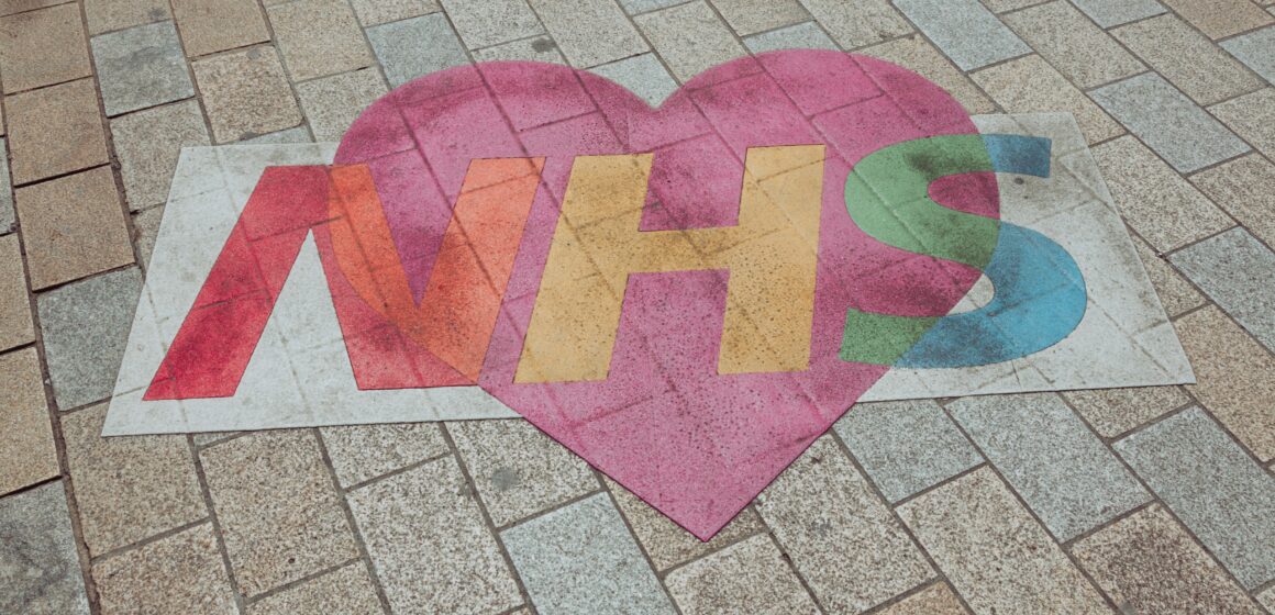nicolas-j-leclercq-fbovpZ4GuLg-unsplash Photograph of the NHS logo painted on the floor with a heart around it.